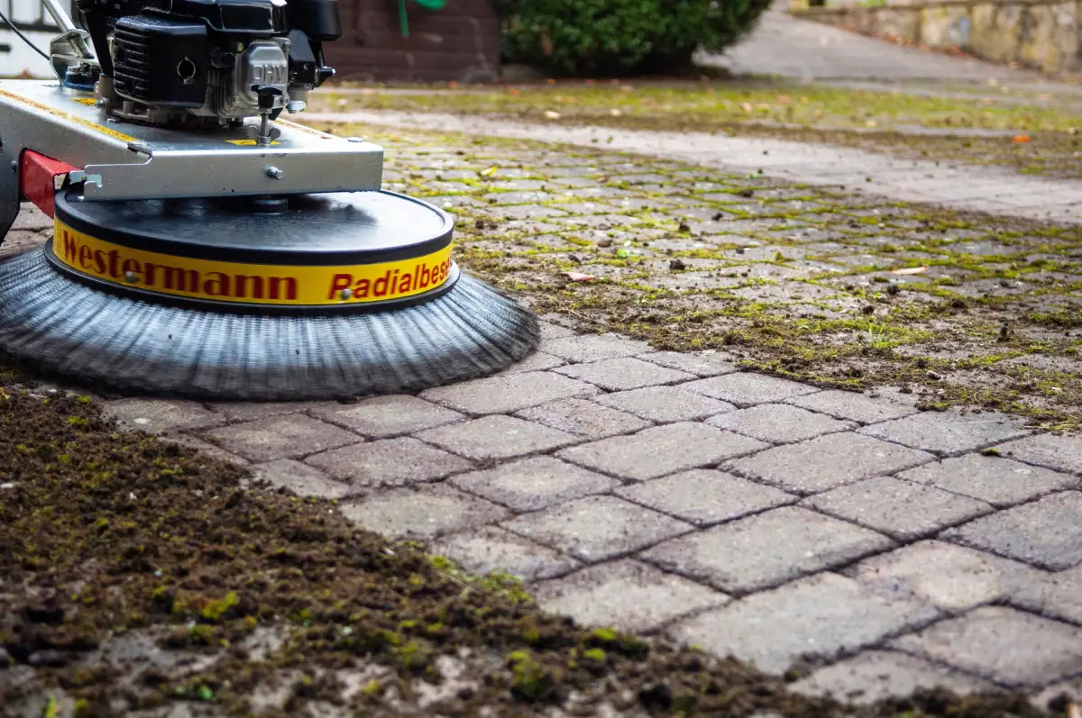 A rotary sweeper lifting moss from paver joints, cleared paving behind it